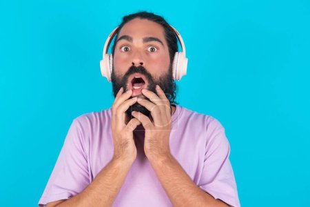 Téléchargez les photos : Choqué homme caucasien avec barbe portant T-shirt violet sur fond bleu regarde fixement la caméra garde la bouche largement ouverte porte des écouteurs stéréo sans fil sur les oreilles - en image libre de droit