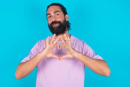 Foto de Hombre caucásico serio con barba vistiendo camiseta violeta sobre fondo azul mantiene las manos cruzadas se levanta en actitud reflexiva concentrado en algún lugar - Imagen libre de derechos