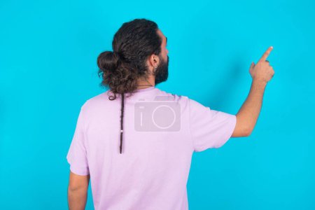 Téléchargez les photos : Homme caucasien avec barbe portant un T-shirt violet sur fond bleu pointant vers l'objet sur l'espace de copie, vue arrière. Tourne le dos - en image libre de droit
