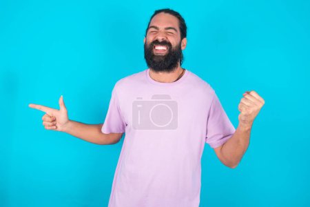 Foto de Alegre hombre caucásico con barba vistiendo camiseta violeta sobre fondo azul mostrando espacio de copia anuncio celebrando suerte - Imagen libre de derechos