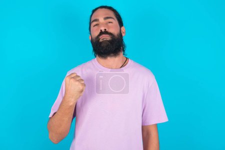 Photo for Caucasian man with beard wearing violet T-shirt over blue background  shows fist has annoyed face expression going to revenge or threaten someone makes serious look. I will show you who is boss - Royalty Free Image