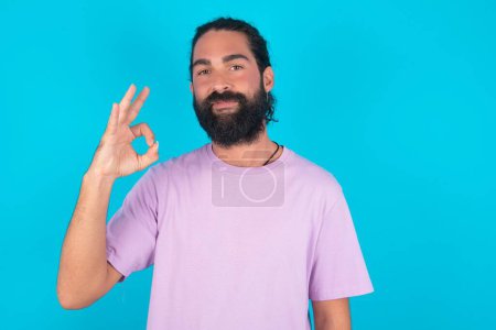 Photo for Caucasian man with beard wearing violet T-shirt over blue background  hold hand arm okey symbol toothy approve advising novelty news - Royalty Free Image