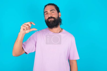 Téléchargez les photos : Homme caucasien mécontent avec barbe portant T-shirt violet sur fond bleu formes petit signe de la main démontre quelque chose de pas très grand. Concept de langage corporel. - en image libre de droit