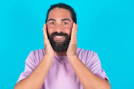 Photo for Happy Caucasian man with beard wearing violet T-shirt over blue background  touches both cheeks gently, has tender smile, shows white teeth, gazes positively straightly at camera, - Royalty Free Image