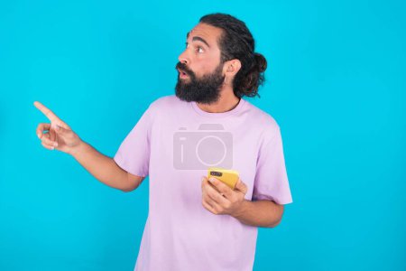 Téléchargez les photos : Homme caucasien étourdi avec barbe portant T-shirt violet sur fond bleu points latéralement droit espace de copie, recommande le produit, voit chose étonnante - en image libre de droit
