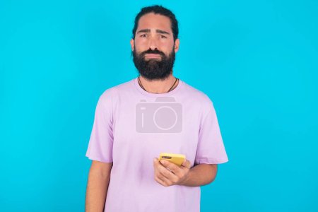 Foto de Hombre caucásico trastornado insatisfecho con barba que usa camiseta violeta sobre fondo azul utiliza aplicación de software móvil y navega información en Internet, sostiene la mano móvil moderna - Imagen libre de derechos