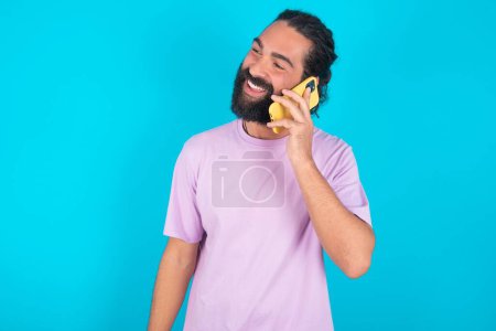 Photo for Pleasant looking happy Caucasian man with beard wearing violet T-shirt over blue background  has nice telephone conversation and looks aside, has nice mood and smiles positively while talks via cell phone - Royalty Free Image