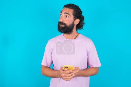 Foto de Hombre caucásico con barba vistiendo camiseta violeta sobre fondo azul sostiene teléfono móvil utiliza Internet de alta velocidad y las redes sociales tiene comunicación en línea. Concepto de tecnologías modernas - Imagen libre de derechos
