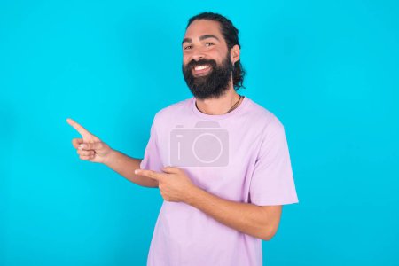 Téléchargez les photos : Homme caucasien avec barbe portant T-shirt violet sur fond bleu points à l'espace de copie indique pour la publicité donne la bonne direction - en image libre de droit