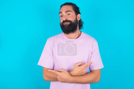 Foto de Hombre caucásico con barba vistiendo camiseta violeta sobre fondo azul cruza brazos y puntos en diferentes lados duda entre dos artículos o variantes. Necesita ayuda con la decisión - Imagen libre de derechos
