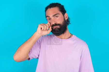 Foto de Decepcionado hombre caucásico abatido con barba vistiendo camiseta violeta sobre fondo azul limpia lágrimas se destaca con expresión sombría. Emoción negativa - Imagen libre de derechos