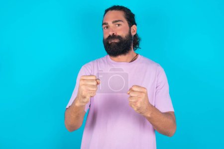 Photo for Displeased annoyed Caucasian man with beard wearing violet T-shirt over blue background  clenches fists, gestures pissed, ready to revenge, looks with aggression at camera stands full of hate, being pressured - Royalty Free Image