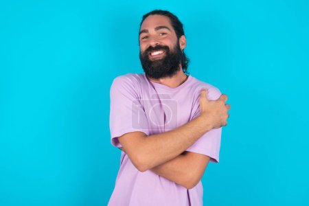 Téléchargez les photos : Charmant homme caucasien heureux avec barbe portant T-shirt violet sur fond bleu embrasse son propre corps, se sent agréablement poses confortables. Concept de tendresse et d'estime de soi - en image libre de droit