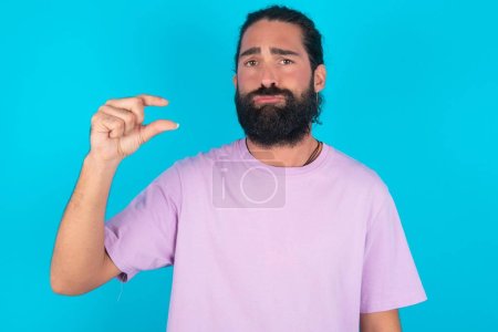 Foto de Hombre caucásico con barba vistiendo camiseta violeta sobre fondo azul monederos labio y gestos con la mano, muestra algo muy poco. - Imagen libre de derechos