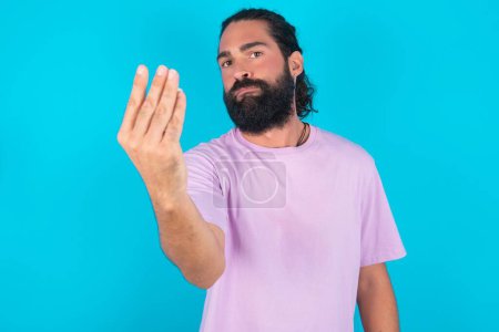 Foto de Hombre caucásico con barba vistiendo camiseta violeta sobre fondo azul haciendo gesto italiano con la mano y los dedos expresión confiada - Imagen libre de derechos