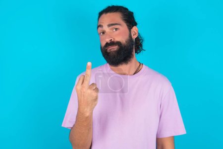 Foto de Hombre caucásico con barba vistiendo camiseta violeta sobre fondo azul muestra dedo medio mala señal pide no molestarse. Provocación y actitud grosera. - Imagen libre de derechos