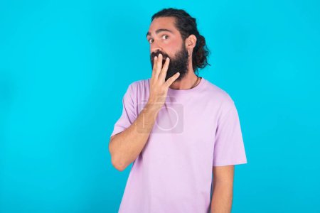 Téléchargez les photos : Homme caucasien avec barbe portant T-shirt violet sur fond bleu couvre la bouche et regarde avec émerveillement à la caméra, ne peut pas croire rumeurs inattendues. - en image libre de droit
