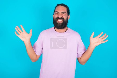 Téléchargez les photos : Homme caucasien fou outré avec barbe portant T-shirt violet sur fond bleu crie bruyamment et des gestes hurlent furieusement. Négatif émotions humaines sentiments concept - en image libre de droit