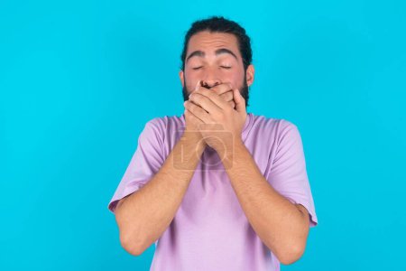 Photo for Stunned Caucasian man with beard wearing violet T-shirt over blue background  covers mouth with both hands being afraid from something or after hearing stunning gossips. - Royalty Free Image