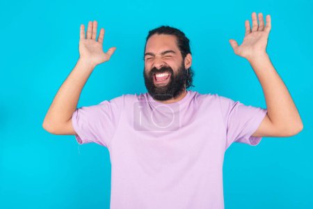 Photo for Caucasian man with beard wearing violet T-shirt over blue  background  goes crazy as head goes around feels stressed because of horrible situation - Royalty Free Image