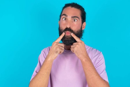 Téléchargez les photos : Homme caucasien avec barbe portant T-shirt violet sur fond bleu croise les yeux et rend les lèvres de poisson grimace drôle - en image libre de droit