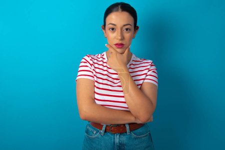 Photo for Thoughtful smiling woman wearing striped T-shirt keeps hand under chin, looks directly at camera, listens something with interest. Youth concept. - Royalty Free Image
