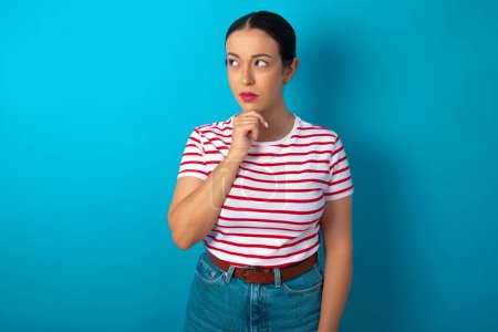 Photo for Shot of contemplative thoughtful woman wearing striped T-shirt keeps hand under chin, looks thoughtfully upwards. - Royalty Free Image
