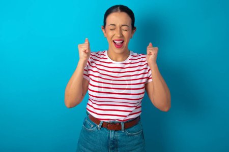 Photo for Woman wearing striped T-shirt rejoicing his success and victory clenching fists with joy being happy to achieve aim and goals. Positive emotions, feelings. - Royalty Free Image
