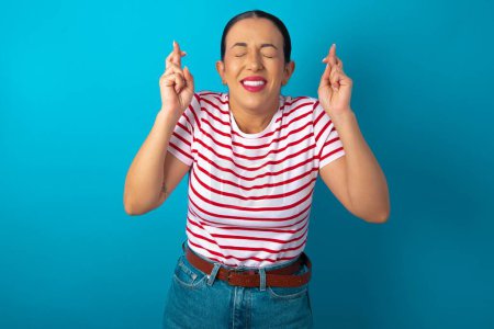 Photo for Joyful woman wearing striped T-shirt clenches teeth, raises fingers crossed, makes desirable wish, waits for good news, I have to win. - Royalty Free Image
