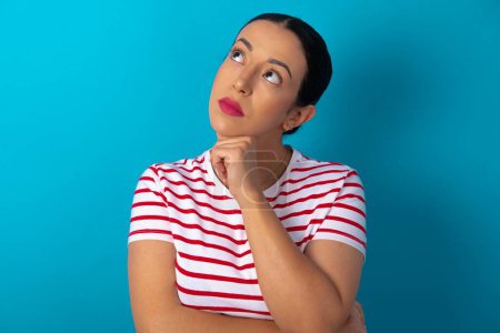 Photo for Portrait of thoughtful woman wearing striped T-shirt keeps hand under chin, looks away trying to remember something or listens something with interest. Youth concept. - Royalty Free Image