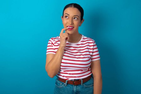 Photo for Woman wearing striped T-shirt with thoughtful expression, looks to the camera, keeps hand near face, bitting a finger thinks about something pleasant. - Royalty Free Image
