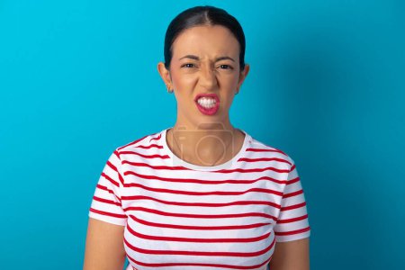 Photo for Mad crazy woman wearing striped T-shirt clenches teeth angrily, being annoyed with coming noise. Negative feeling concept. - Royalty Free Image