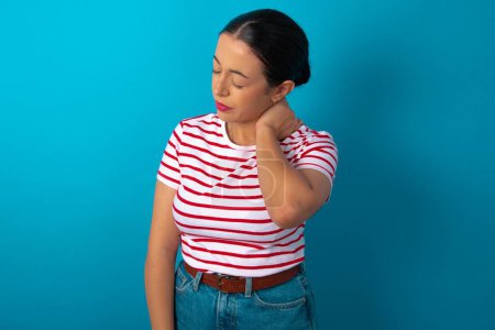 Foto de Mujer con camiseta rayada que sufre de lesión en la espalda y el cuello, tocando el cuello con la mano, dolor muscular. - Imagen libre de derechos