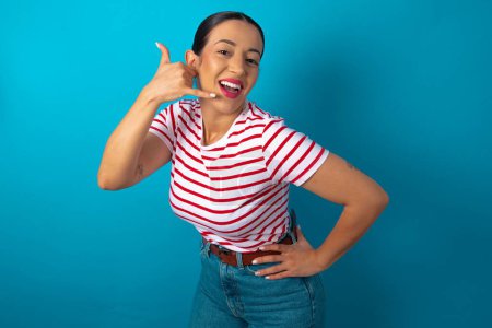 Photo for Woman wearing striped T-shirt imitates telephone conversation, makes phone call gesture with hands, has confident expression. Call me! - Royalty Free Image