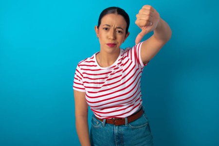 Photo for Discontent woman wearing striped T-shirt shows disapproval sign, keeps thumb down, expresses dislike, frowns face in discontent. Negative feelings. - Royalty Free Image