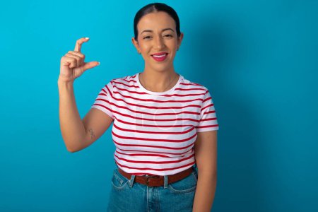 Foto de Mujer vistiendo camiseta a rayas sonriendo y haciendo gestos con la mano de tamaño pequeño, símbolo de medida - Imagen libre de derechos