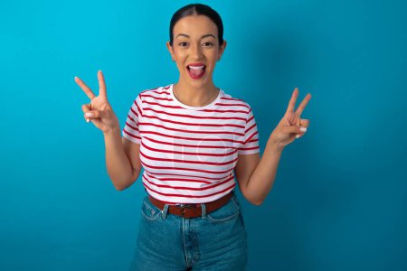Photo for Woman wearing striped T-shirt with optimistic smile, showing peace or victory gesture with both hands, looking friendly. V sign. - Royalty Free Image