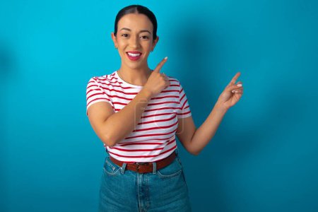 Photo for Positive woman wearing striped T-shirt with beaming smile pointing with two fingers and looking on empty copy space. Advertisement concept. - Royalty Free Image
