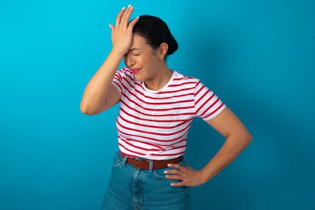 Foto de Mujer frustrada vistiendo una camiseta a rayas cogida de la mano en la frente deprimida lamentando lo que hizo con dolor de cabeza, luciendo estresante. - Imagen libre de derechos
