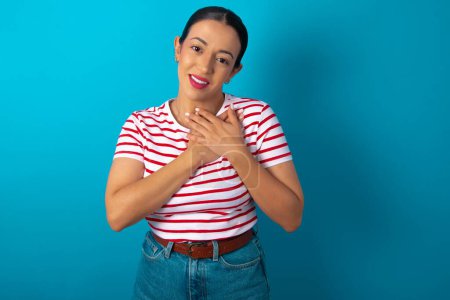 Photo for Honest woman wearing striped T-shirt keeps hands on chest, touched by compliment or makes promise, looks at camera with great pleasure. - Royalty Free Image