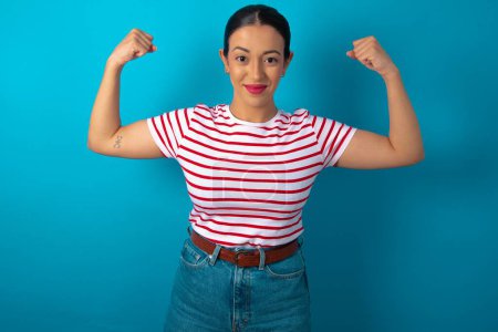 Photo for Waist up shot of woman wearing striped T-shirt raises arms to show muscles feels confident in victory, looks strong and independent, smiles positively at camera. Sport concept. - Royalty Free Image