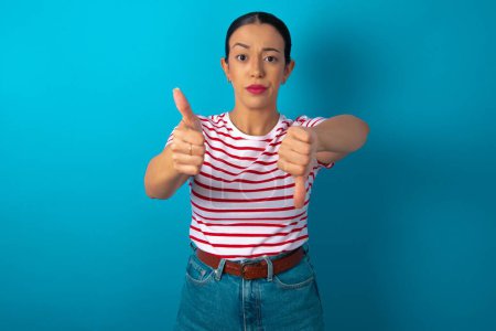 Photo for Woman wearing striped T-shirt feeling unsure making good bad sign. Displeased and unimpressed. - Royalty Free Image