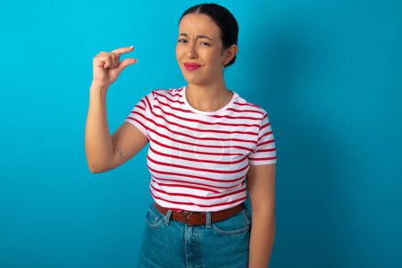 Photo for Upset woman wearing striped T-shirt shapes little gesture with hand demonstrates something very tiny small size. Not very much - Royalty Free Image