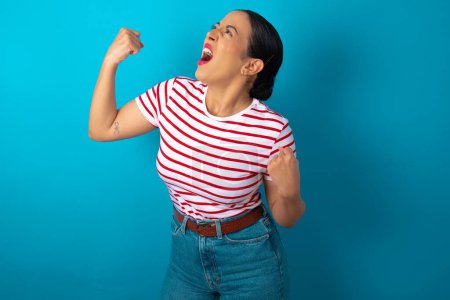 Photo for Attractive woman wearing striped T-shirt celebrating a victory punching the air with his fists and a beaming toothy smile - Royalty Free Image