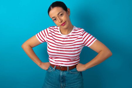 Photo for Funny frustrated woman wearing striped T-shirt holding hands on waist and silly looking at awkward situation. - Royalty Free Image