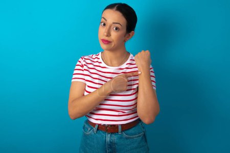 Téléchargez les photos : Femme portant des rayures T-shirt à la hâte pointant pour regarder le temps, l'impatience, bouleversé et en colère pour retard de délai - en image libre de droit