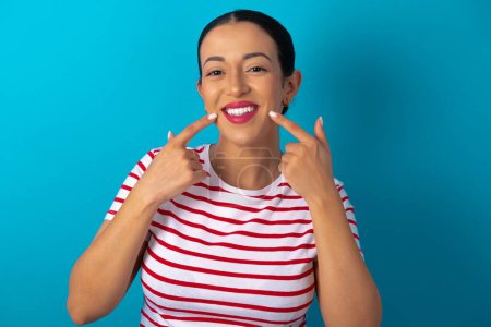 Téléchargez les photos : Dents blanches droites saines fortes. Portrait rapproché d'une femme heureuse portant un T-shirt rayé avec un sourire rayonnant pointant sur des dents blanches claires et parfaites. - en image libre de droit