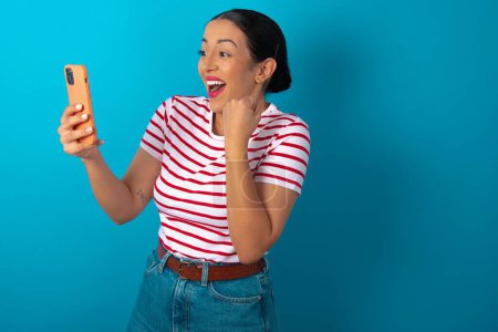 Photo for Happy cheerful woman wearing striped T-shirt receiving good news via e-mail and celebrating success while standing and looking at mobile phone. - Royalty Free Image