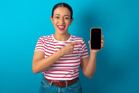 Photo for Smiling woman wearing striped T-shirt showing and pointing at empty phone screen. Advertisement and communication concept. - Royalty Free Image
