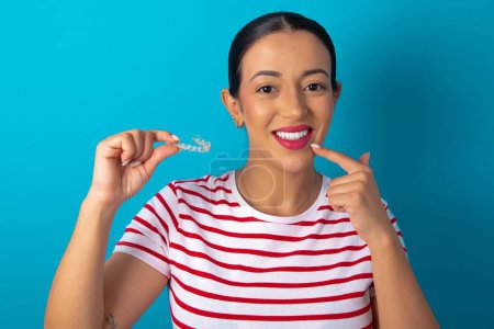 mujer con camiseta a rayas sosteniendo un alineador invisible y señalando sus dientes rectos perfectos. Salud dental y concepto de confianza.